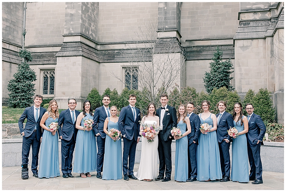 A photo of a bride and groom with their large wedding party, dressed in dusty blue dresses and tuxes holding pastel spring florals | Atlanta pastel church wedding inspiration